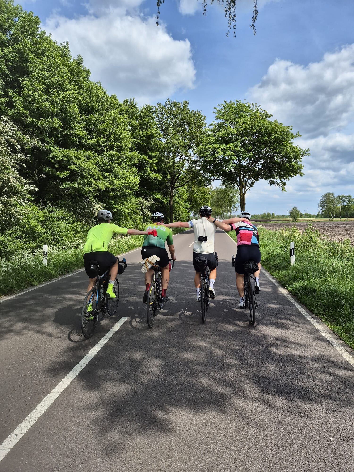 Four cyclists riding side-by-side with arms linked on a country road