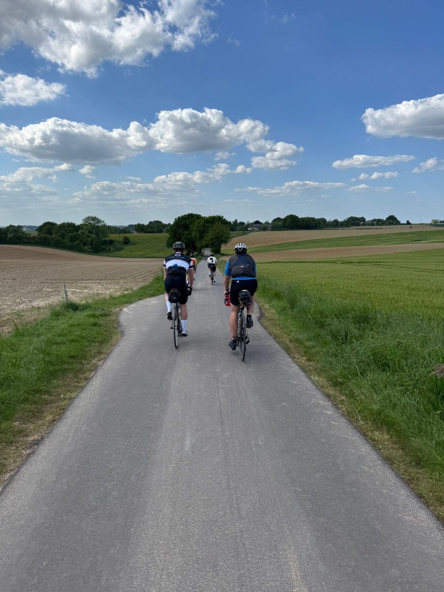 Three cyclists riding a quiet country lane through rolling Ardennes fields under cumulus clouds
