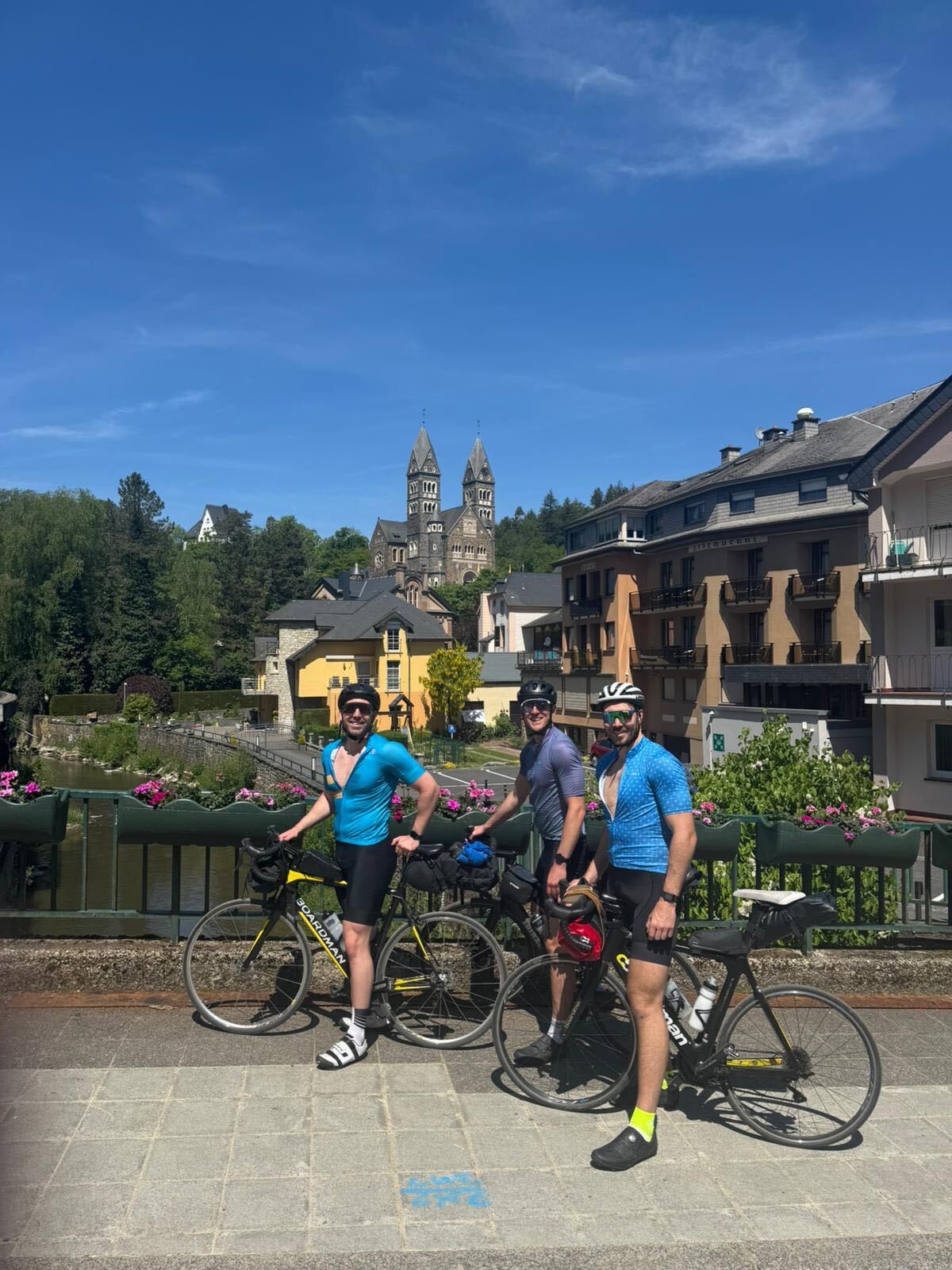 Three cyclists posing on a bridge with a twin-spired Luxembourg church on the hillside behind