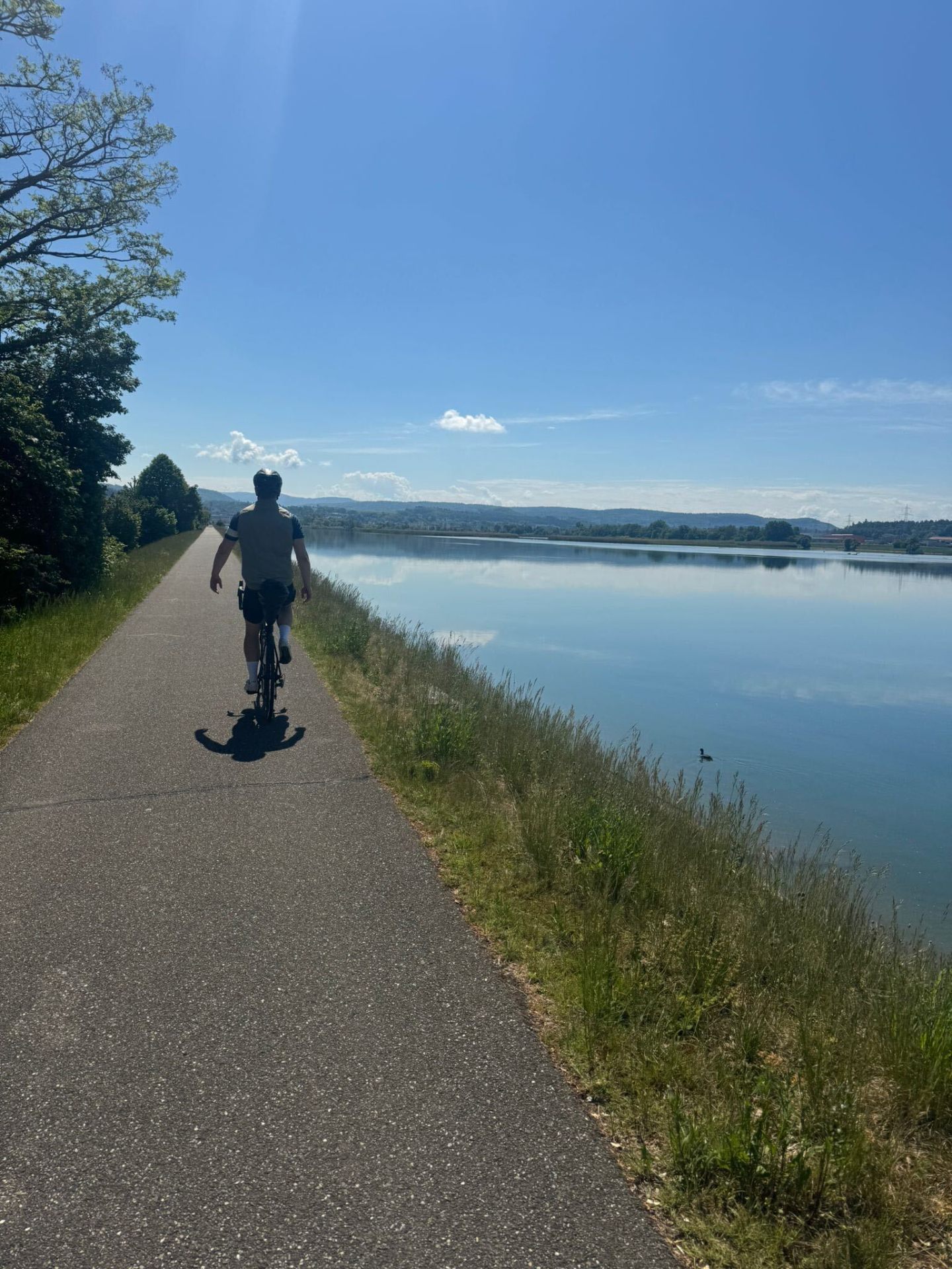 Lone rider on a lakeside path with mirror-calm water and distant Swiss hills